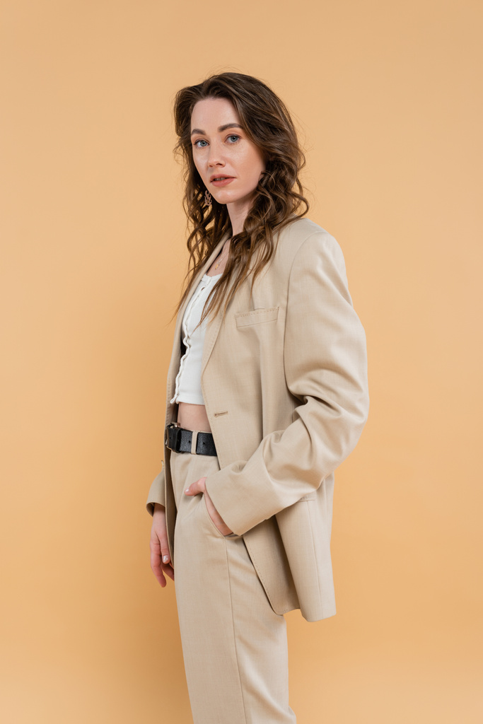 style and fashion concept, young woman with wavy hair standing in fashionable suit while posing on beige background, formal attire, hand in pocket, looking at camera, modern elegance  - Photo, Image