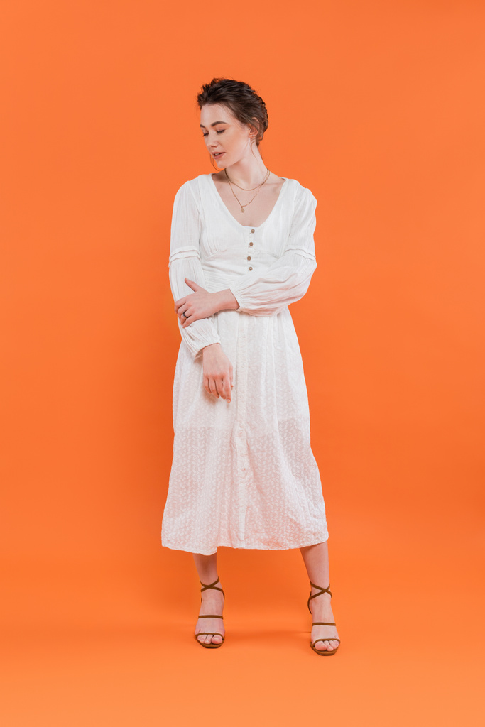 літня мода, молода жінка в білому сонячному платті, дивлячись вниз і стоячи на помаранчевому фоні, яскравий фон, стильна позування, леді в білому, модний тренд, елегантність
  - Фото, зображення