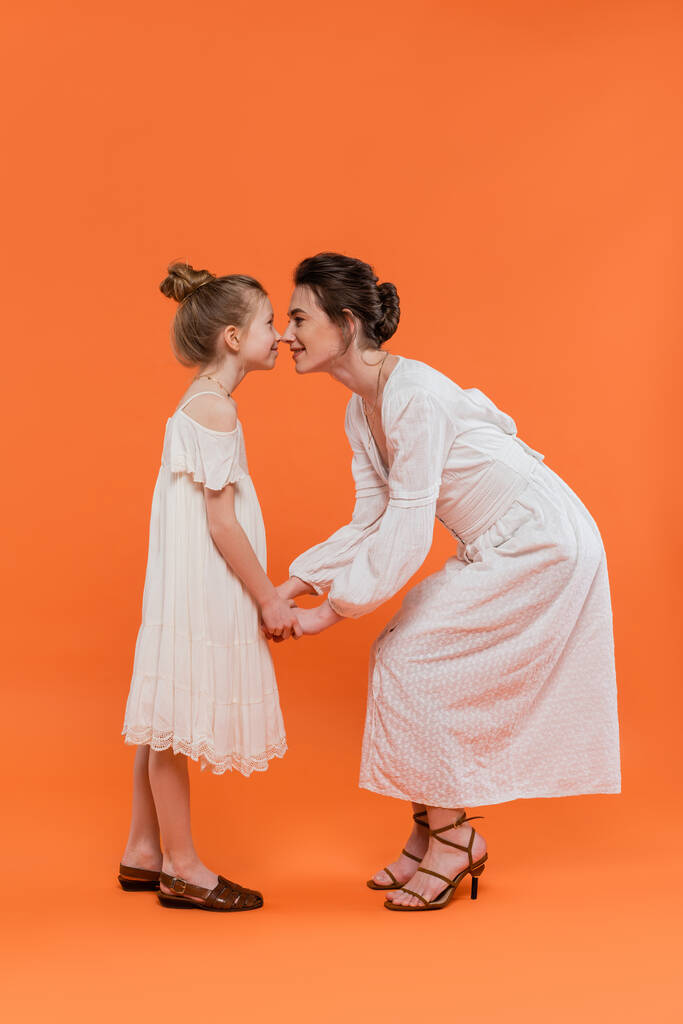 summer trends, young mother holding hands with happy preteen daughter and standing on orange background, white sun dresses, togetherness, fashion and style concept, bonding and love, nose to nose - Photo, Image