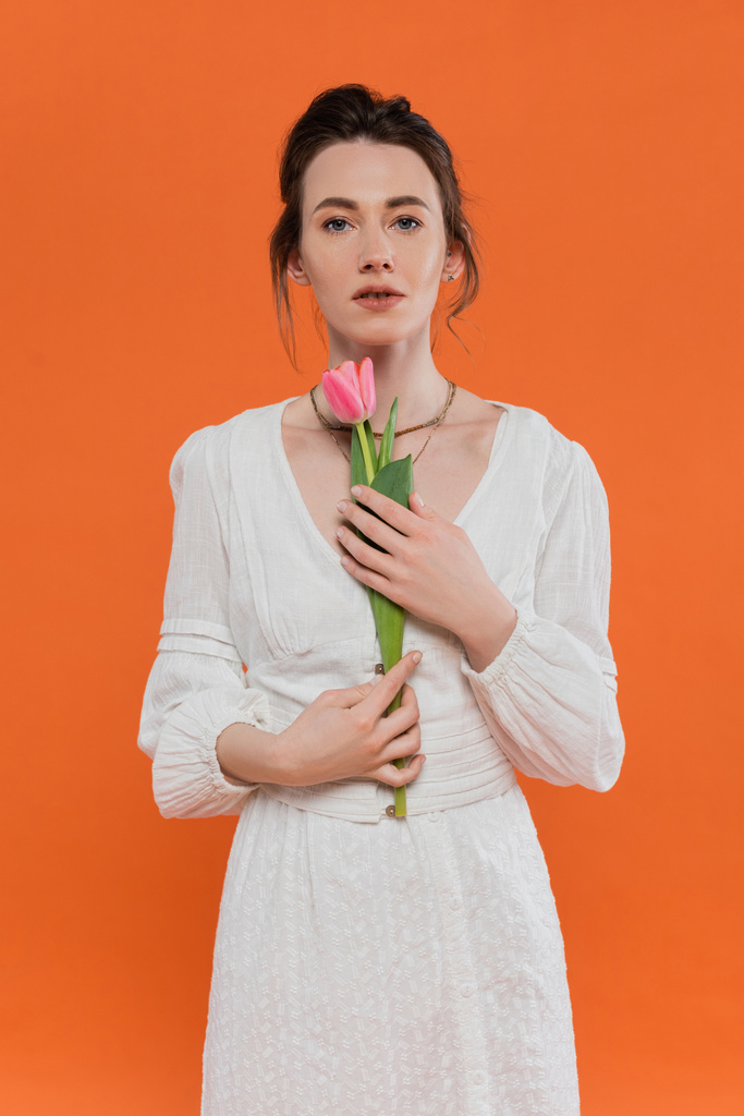 smyslnost, každodenní móda, mladá žena v bílých slunečních šatech drží růžový tulipán a stojí na oranžovém pozadí, dáma v bílém, pulzující pozadí, móda a příroda, léto, portrét  - Fotografie, Obrázek