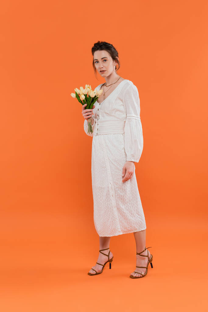 concept de mode, jolie jeune femme en robe de soleil blanche tenant des tulipes jaunes et debout sur fond orange, dame en blanc, fond vibrant, mode, été, bouquet de fleurs, pleine longueur - Photo, image