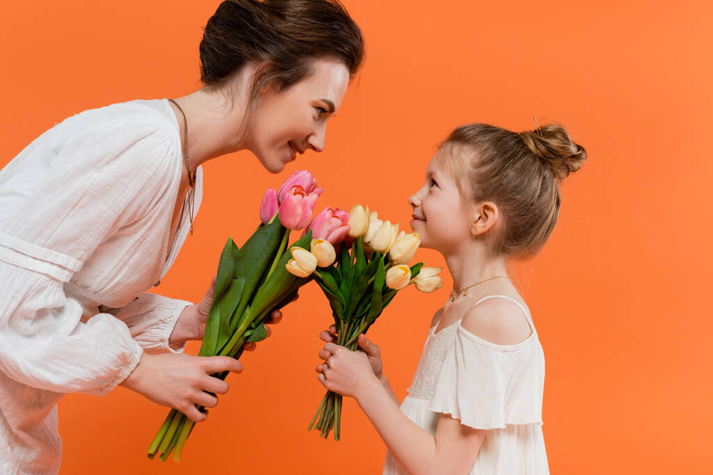 ευτυχισμένη μητέρα και παιδί με λουλούδια, νεαρή γυναίκα και κορίτσι κρατώντας τουλίπες και κοιτάζοντας ο ένας τον άλλον σε πορτοκαλί φόντο, καλοκαιρινή μόδα, φορέματα ήλιο, θηλυκό δέσιμο  - Φωτογραφία, εικόνα