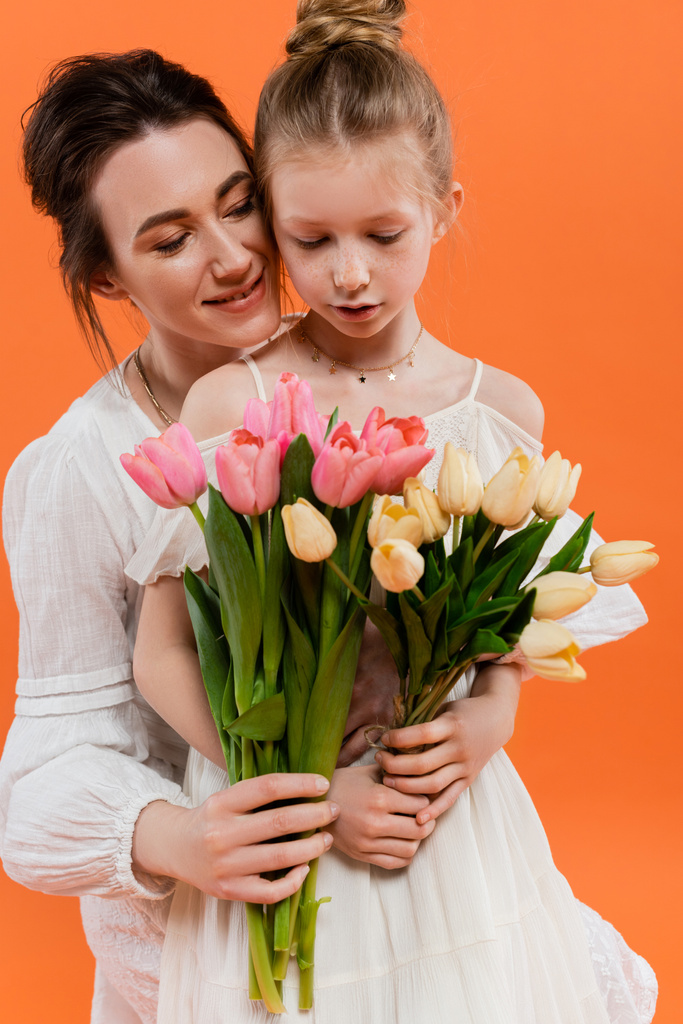 happy mother and daughter with tulips, young woman and girl holding flowers and posing on orange background, summer fashion,  family love, sun dresses, female bonding  - Photo, Image