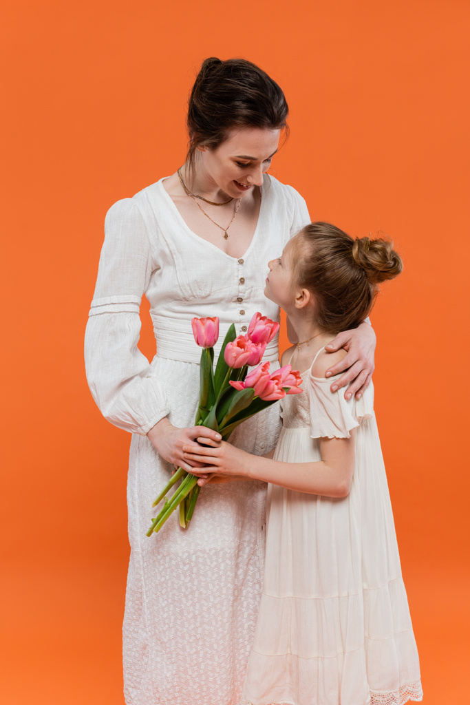 Dzień matki, matka przytulająca nastoletnią córkę z bukietem kwiatów na pomarańczowym tle, wiązanie, białe sukienki, różowe tulipany, szczęśliwe wakacje, żywe kolory, radosna okazja  - Zdjęcie, obraz