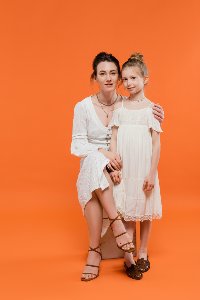 motherly love, brunette young woman sitting on chair and hugging preteen daughter on orange background, white sun dresses, summer fashion, togetherness, love, female bonding, modern parenting  - Photo, Image