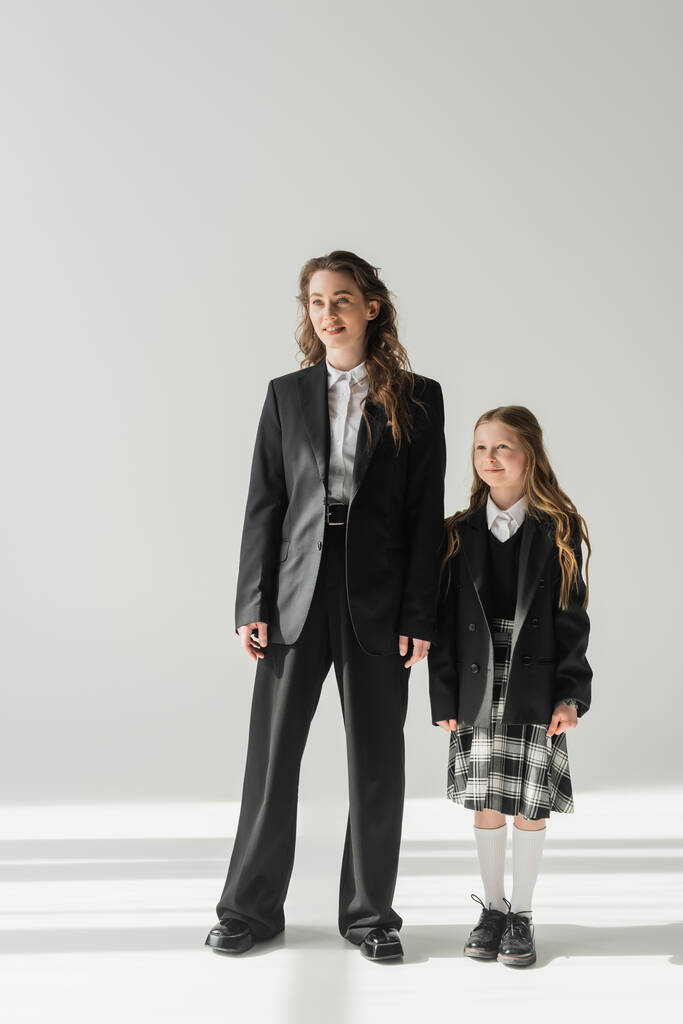 εργαζόμενη μητέρα και μαθήτρια, χαρούμενο κορίτσι με σχολική στολή στέκεται με την επιχειρηματίας με κοστούμι σε γκρι φόντο, κρατώντας τα χέρια, επίσημη ενδυμασία, μοντέρνα οικογένεια, δέσιμο, σύγχρονη ανατροφή των παιδιών  - Φωτογραφία, εικόνα