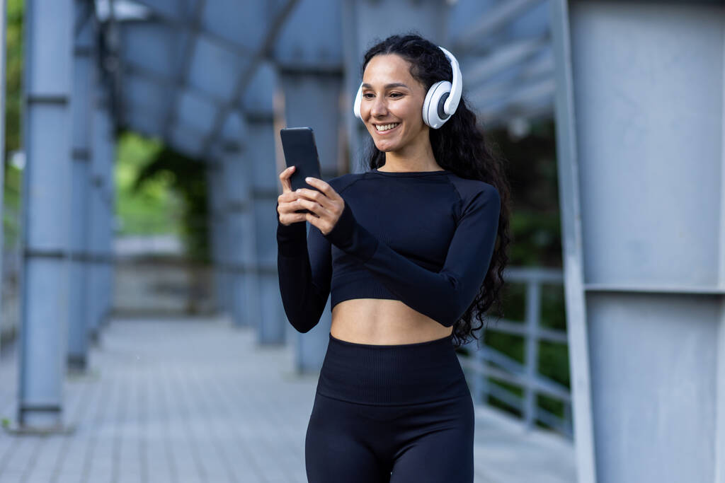 Deportista hispana con auriculares escuchando música en línea y podcasts de audio con libros mientras trota y hace ejercicios activos, mujer usando aplicación en el teléfono, en chándal. - Foto, Imagen
