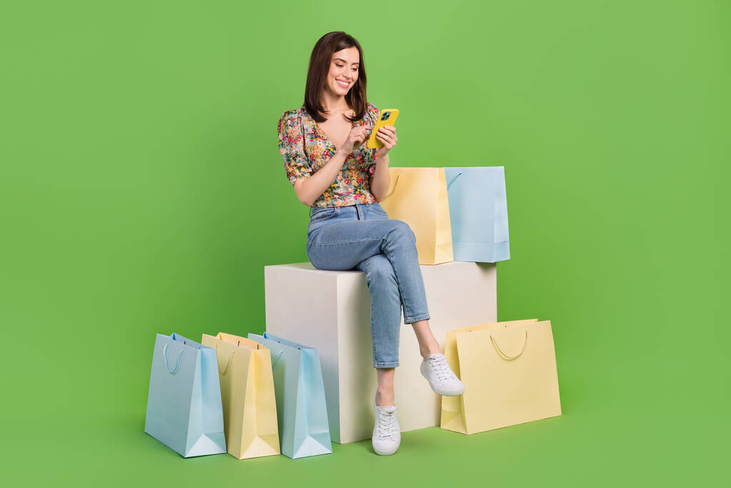 Pełna długość zdjęcie uroczej osoby siedzieć platforma sześcian używać smartfona zamówić torby centrum handlowego izolowane na zielonym tle kolor. - Zdjęcie, obraz
