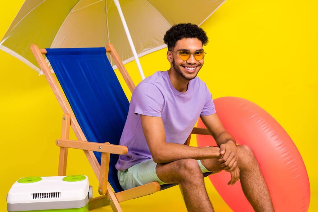 Foto de joven alegre sentado tumbona disfrutar de sus vacaciones fin de semana de verano temporada de verano cerca de la nevera aislado en el fondo de color amarillo. - Foto, imagen