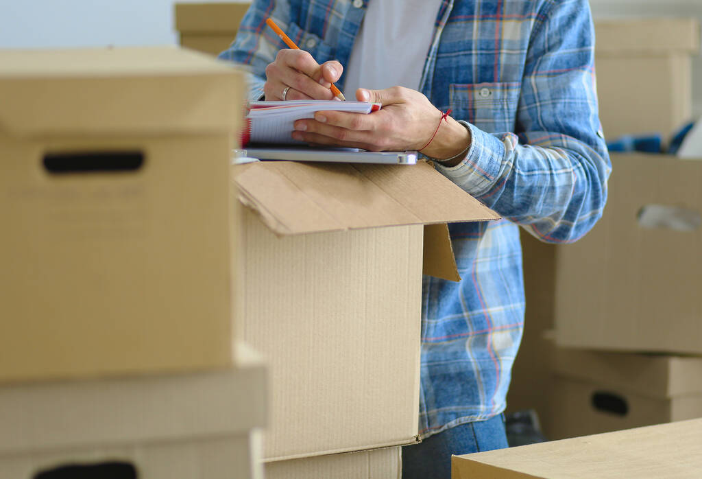 Ένας άνθρωπος που κινείται κάθεται στο πάτωμα σε άδειο διαμέρισμα, ανάμεσα στα κουτιά, ελέγχοντας τη λίστα των πραγμάτων - Φωτογραφία, εικόνα