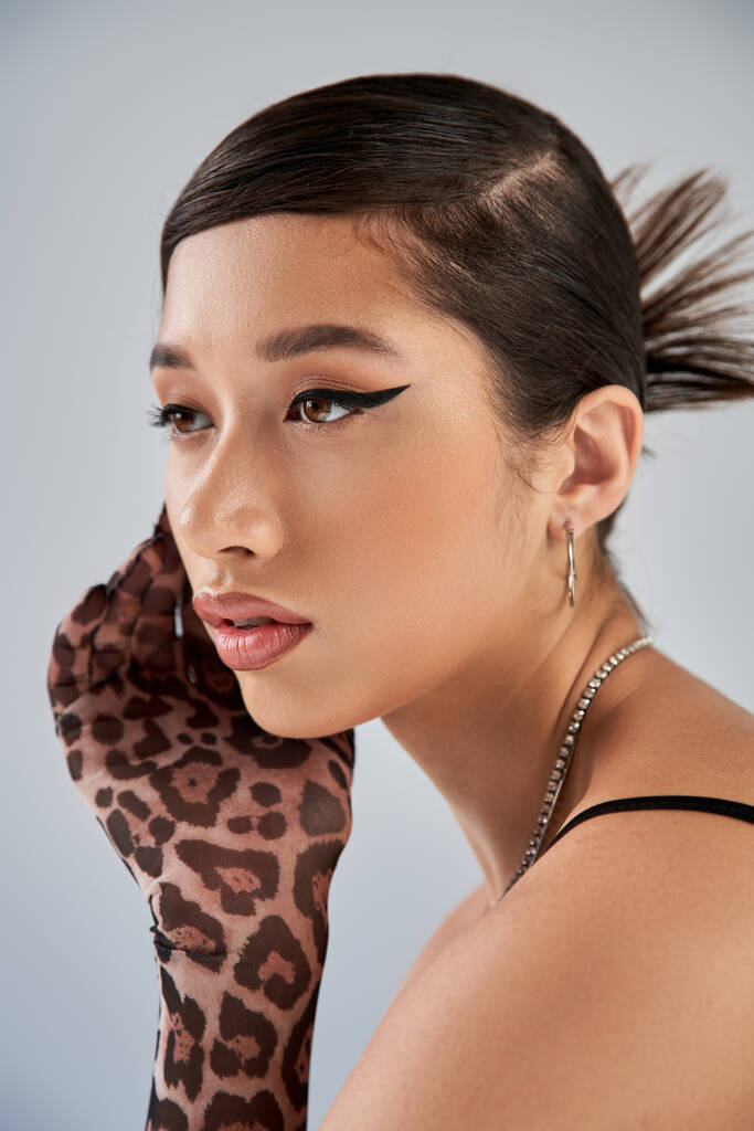 portret van betoverende aziatische vrouw met brunette haar, vet make-up, trendy kapsel, zilveren kettingen en dierlijke print handschoen poseren met de hand in de buurt van gezicht op grijze achtergrond, lente mode - Foto, afbeelding