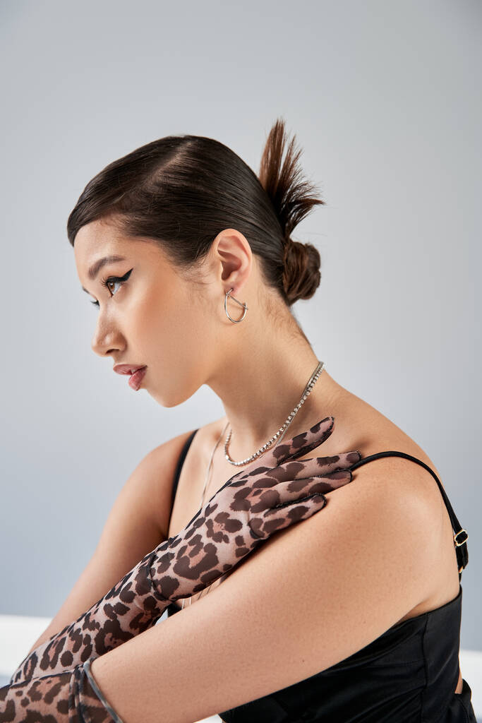 portrait of young asian woman with trendy hairstyle and bold makeup posing in animal print gloves, black strap dress and silver accessories on grey background, spring fashion concept - Photo, Image