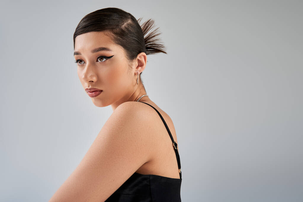 spring fashion photography, portrait of stylish asian model in black strap dress, with trendy hairstyle, bold makeup and expressive gaze looking away on grey background, generation z - Photo, Image