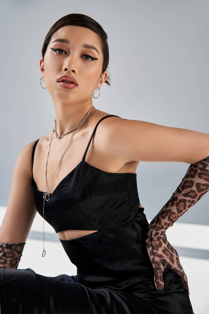 trendy lente, portret van sensuele aziatische vrouw met expressieve blik en heldere make-up, in zwarte bandjurk en zilveren accessoires, hand in hand op heup op grijze achtergrond met verlichting en schaduwen - Foto, afbeelding