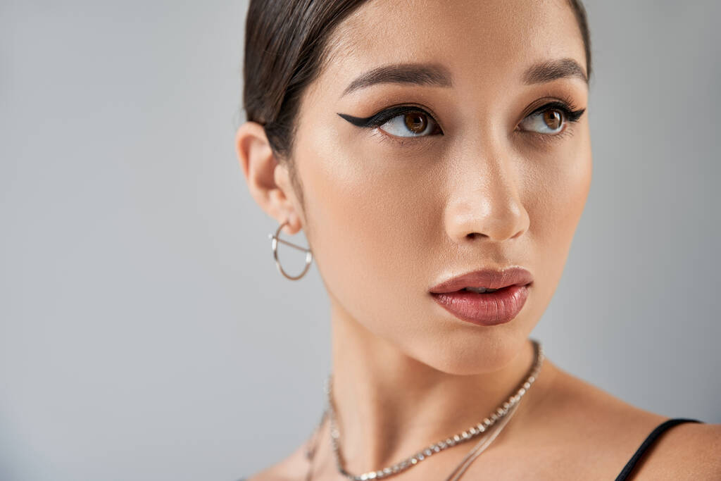 Portret Azjatki w srebrnych naszyjnikach i kolczykach, z brunetką, odważnym makijażem i wyrazistym spojrzeniem odwracającym wzrok na szarym tle, modna wiosna, fotografia mody - Zdjęcie, obraz