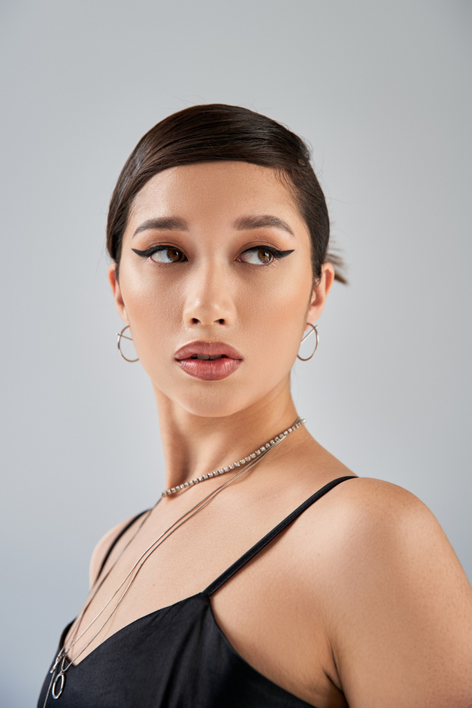 portré csábító ázsiai nő elegáns fekete ruhában, ezüst nyakláncok és fülbevalók, merész smink és barna haj néz félre szürke háttér, divatos tavasz - Fotó, kép