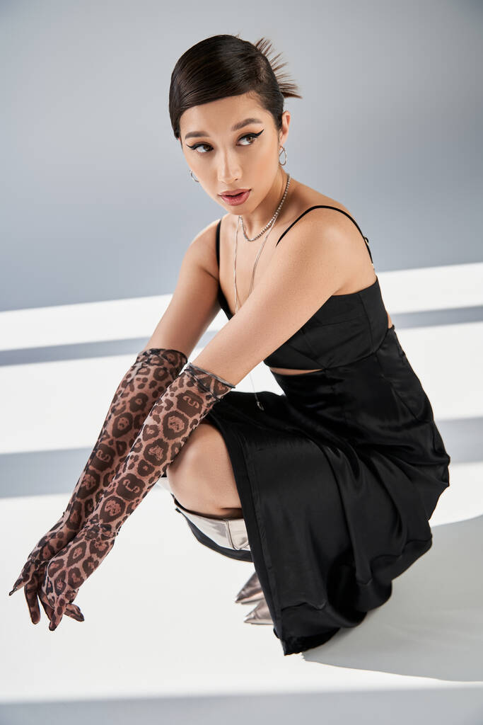 full length of young asian woman in black elegant dress, animal print gloves and silver boots posing on haunches and looking away on grey background with lighting, high fashion, spring style - Photo, Image