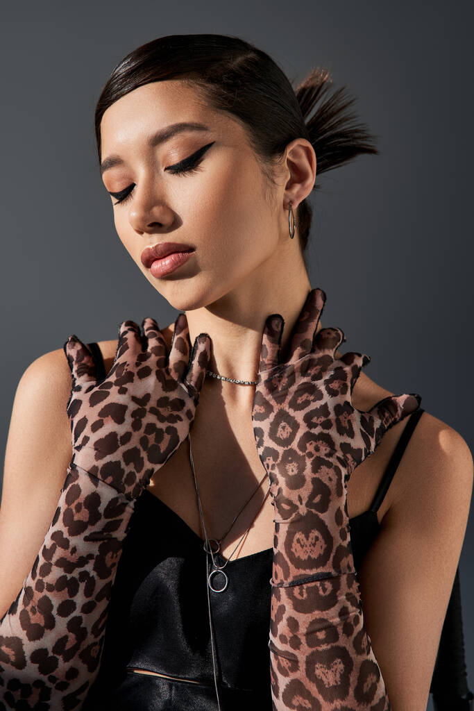 portret van sensuele aziatische vrouw met gedurfde make-up, in zwarte bandjurk en dierenprint handschoenen poseren met gesloten ogen op donkergrijze achtergrond, trendy lenteconcept, generatie z - Foto, afbeelding