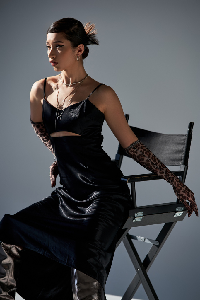jeune femme asiatique avec un maquillage audacieux et coiffure à la mode, en robe élégante noire, colliers en argent et gants d'impression animale posant sur chaise sur fond gris avec éclairage, printemps élégant - Photo, image