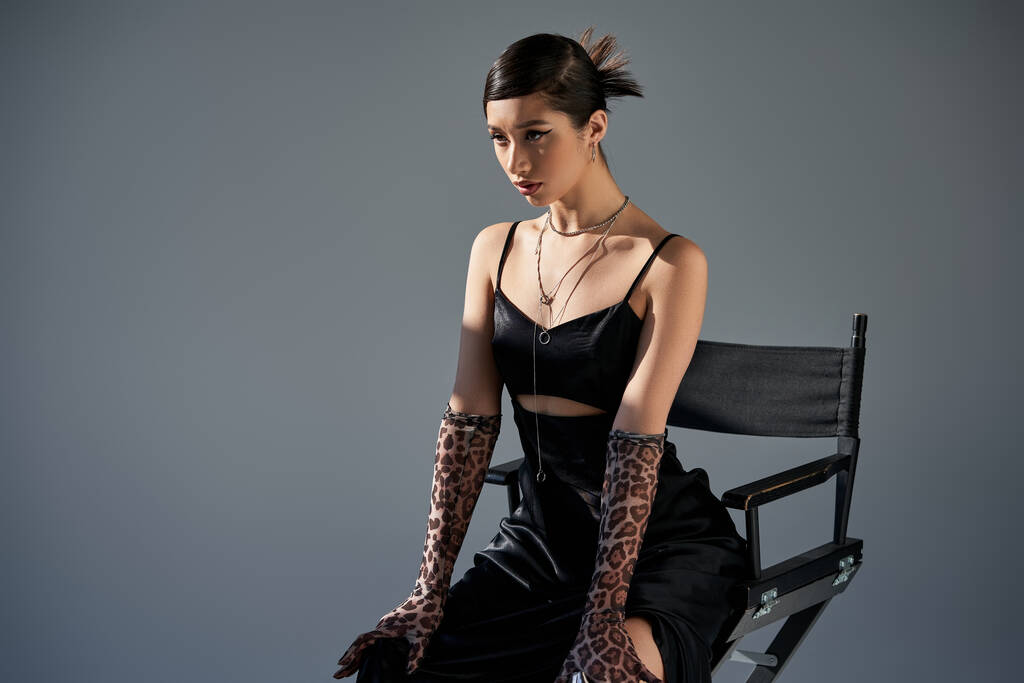 νεαρό ασιατικό μοντέλο μόδας με καστανά μαλλιά ποζάρουν σε καρέκλα σε μοντέρνο φόρεμα άνοιξη, μαύρο λουρί φόρεμα, γάντια animal print, ασημένια κολιέ σε γκρι φόντο με φωτισμό, γενιά z - Φωτογραφία, εικόνα