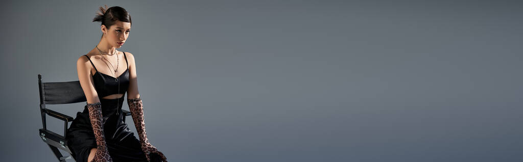 primavera alla moda, donna asiatica espressiva con capelli castani seduta sulla sedia in abito cinturino nero, guanti stampa animalier e guardando lontano su sfondo grigio con illuminazione, fotografia di moda, banner - Foto, immagini