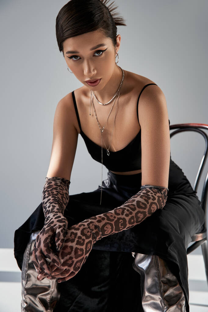 spring fashion photography, asian woman with expressive gaze, in animal print gloves, black strap dress and silver necklaces sitting on chair and looking at camera on grey background with lighting - Photo, Image