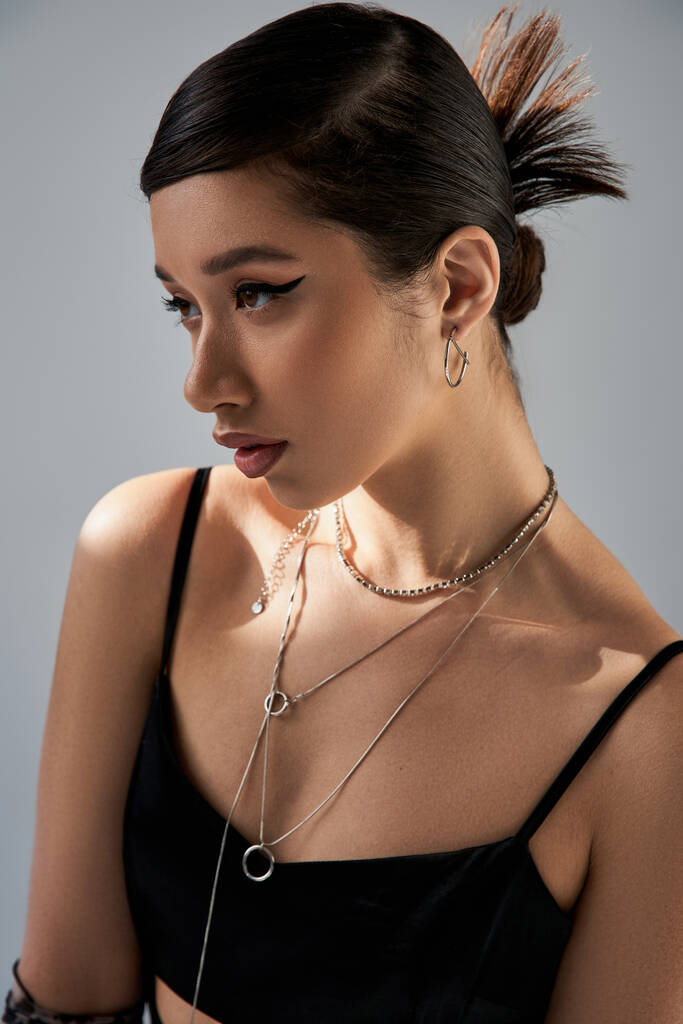 portrait of brunette asian woman with bold makeup, trendy hairstyle and expressive gaze, in black strap dress and silver necklaces looking away on grey background with lighting, trendy spring concept - Photo, Image