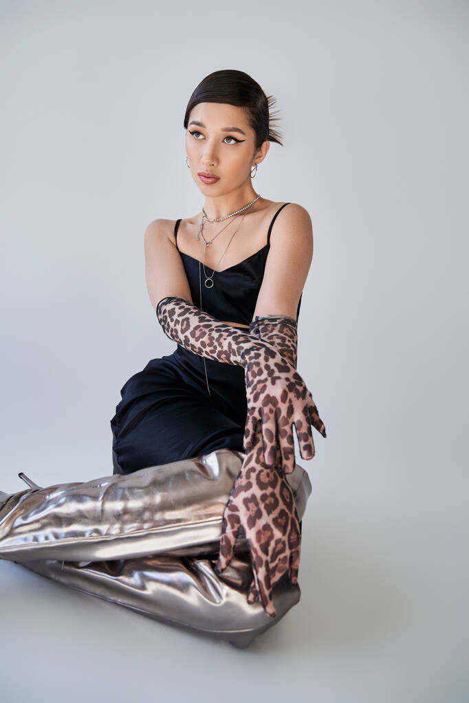 elegant asian woman with bold makeup, in black strap dress, animal print gloves and silver boots sitting and looking away on grey background, youthful style, trendy spring concept - Photo, Image