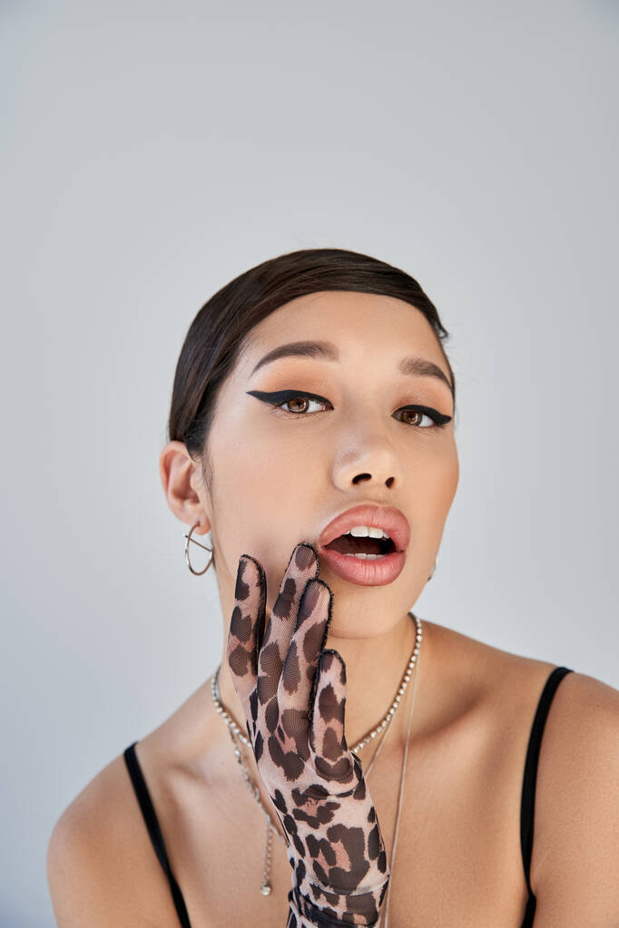 portret uwodzicielskiej azjatki z odważnym makijażem, w srebrnych naszyjnikach i rękawiczkach z nadrukiem zwierzęcia trzymającej się za rękę przy otwartych ustach i patrzącej na kamerę na szarym tle, wiosenna koncepcja mody - Zdjęcie, obraz