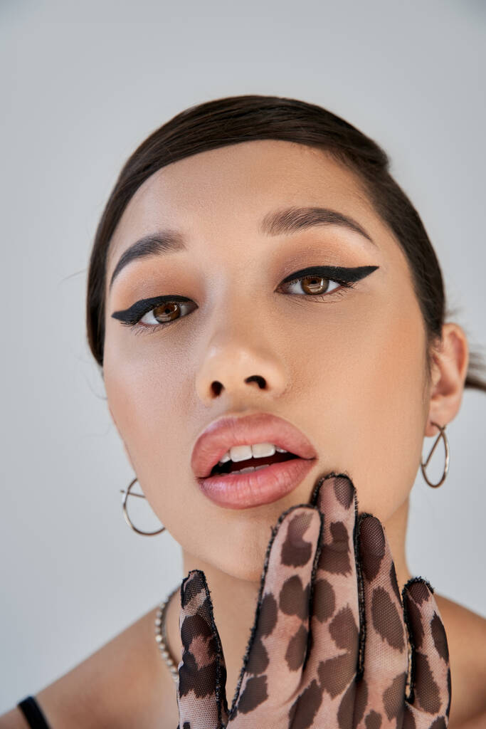 close up portrait of charming asian woman with seductive gaze holding hand near open mouth and looking at camera on grey background, bold makeup, animal print glove, trendy spring concept - Photo, Image