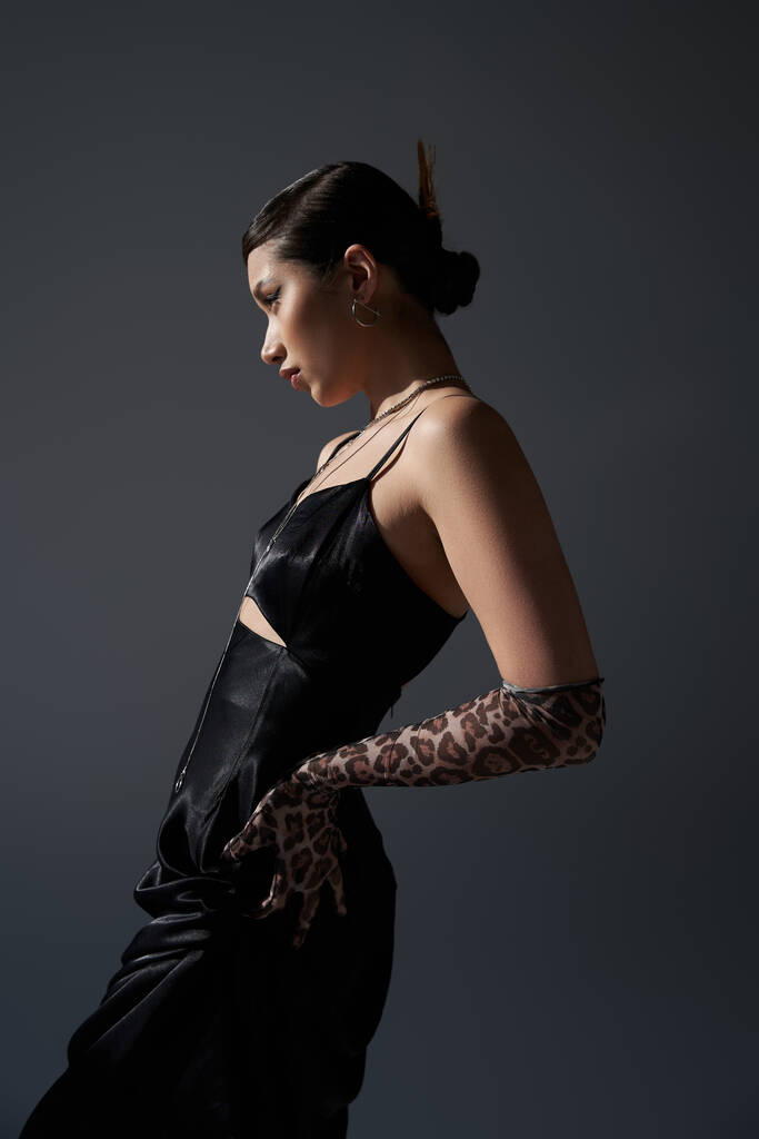 side view of young and pretty asian woman with trendy hairstyle posing with hand on hip in black strap dress and animal print glove on dark grey background, fashionable spring concept - Photo, Image