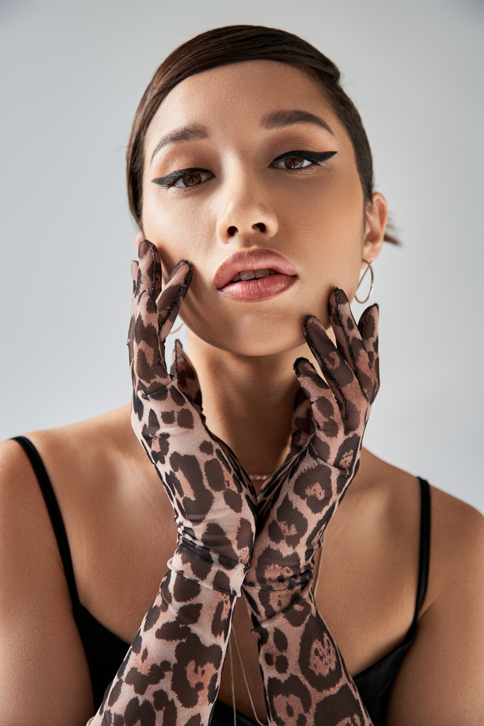 portret van jeugdige en mooie Aziatische mode model met brunette haar en gedurfde make-up poseren in dierlijke print handschoenen en kijken naar de camera op grijze achtergrond, trendy lente concept - Foto, afbeelding