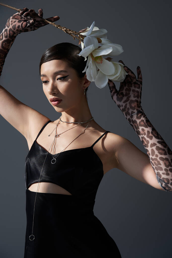 jeugdige aziatische vrouw in zwart elegante bandjurk, zilveren kettingen en dierenprint handschoenen met witte bloeiende orchidee boven het hoofd op donkergrijze achtergrond, trendy lente concept, fashion shoot - Foto, afbeelding