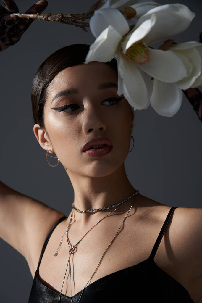 portrait of expressive asian woman in silver necklaces and black strap dress, with brunette hair and bold makeup holding white orchid on dark grey background, fashionable spring concept - Photo, Image