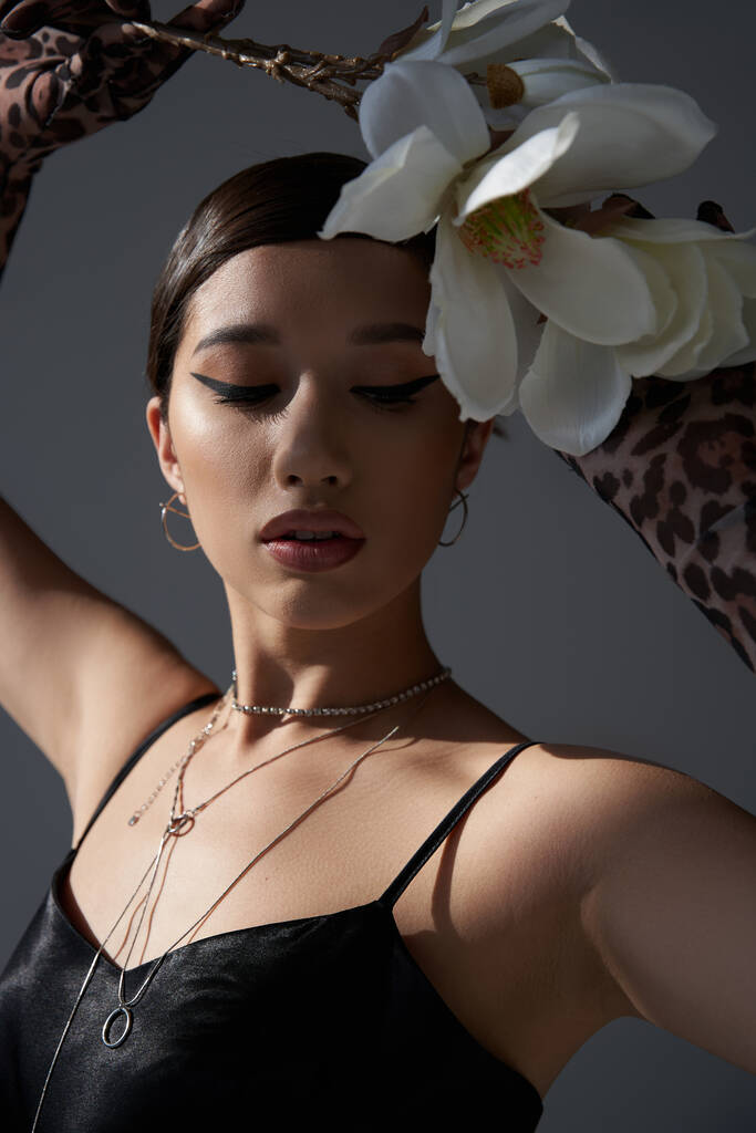 portré fiatal ázsiai divat modell merész smink és barna haj visel fekete pánt ruha és ezüst nyakláncok pózol fehér orchidea sötét szürke háttér, tavaszi stílus koncepció - Fotó, kép