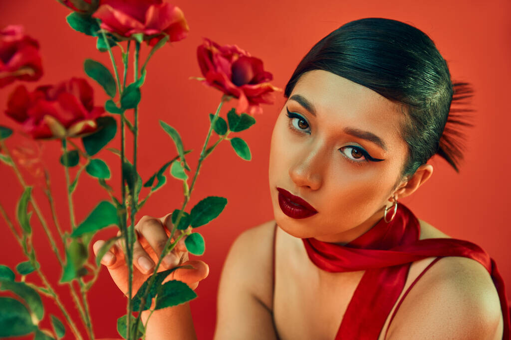 kevät tyyli valokuvaus, viehättävä aasialainen nainen ruskeaverikkö hiukset ja rohkea meikki koskettaa vihreät lehdet ruusut ja katsot kameran punaisella taustalla, sukupolvi z - Valokuva, kuva