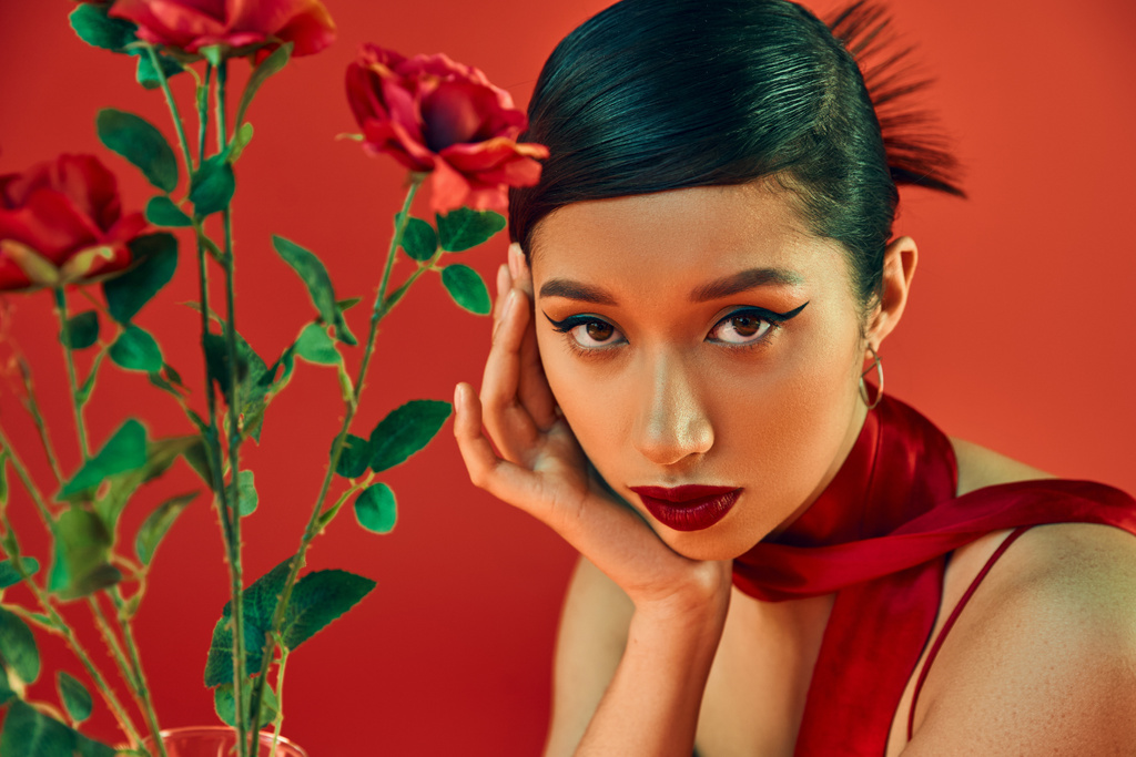 portrait of attractive asian woman with bold makeup, brunette hair and expressive gaze looking at camera near roses on red background, fashionable spring, youthful style - Photo, Image