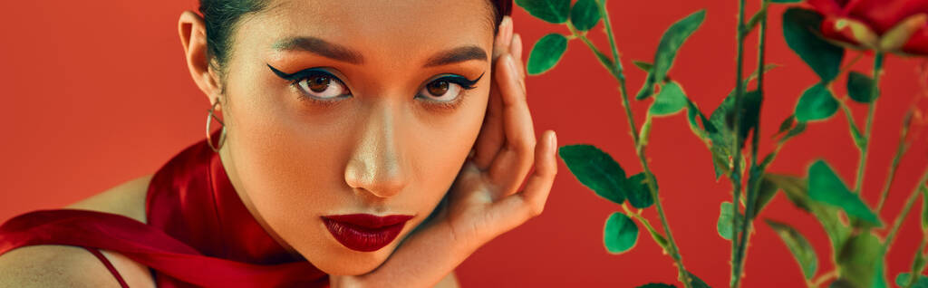portret van jeugdige en verleidelijke Aziatische vrouw met gedurfde make-up en expressieve blik hand in hand in de buurt van gezicht en kijken naar camera in de buurt van bloemen op rode achtergrond, trendy lente, generatie z, banner - Foto, afbeelding