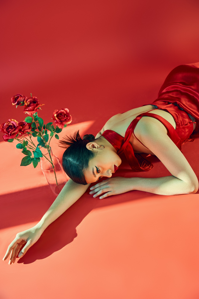 vue grand angle de jeune femme asiatique sensuelle en robe élégante et foulard couché dans l'éclairage sur fond rouge près de vase en verre avec des roses, gen z fashion, concept de printemps à la mode - Photo, image