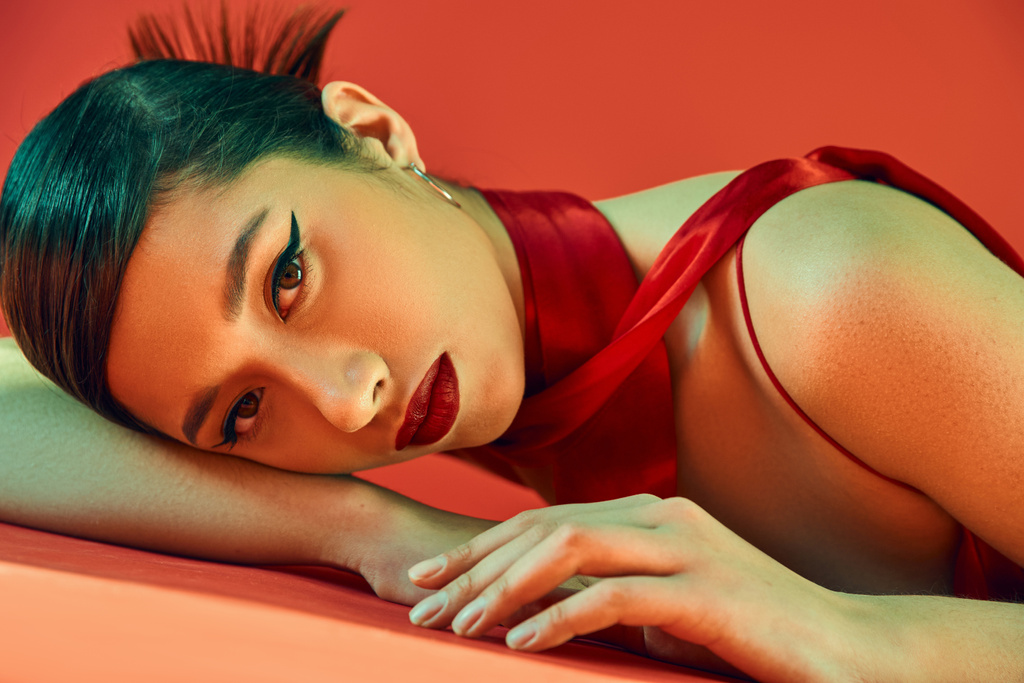 smyslná a okouzlující asijská žena s expresivním pohledem ležící na červeném pozadí a dívající se do kamery, tučný make-up, brunetky vlasy, krční šátek, jarní módní fotografie - Fotografie, Obrázek