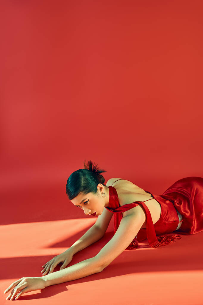 smyslná asijská žena s módním účesem a tučným make-upem, v krčníku a elegantní šaty pózující na červeném pozadí s osvětlením, módní jarní koncept, generace z - Fotografie, Obrázek