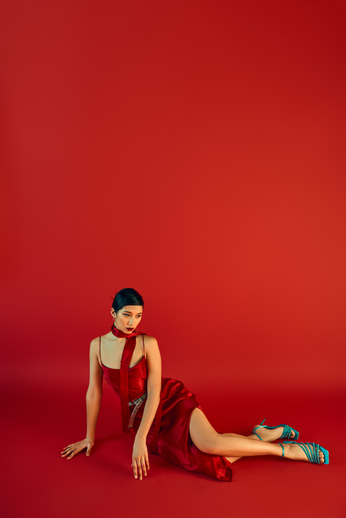 volle Länge der jungen asiatischen Frau mit brünetten Haaren und kühnem Make-up, in elegantem Kleid und Halstuch sitzt auf rotem Hintergrund mit Kopierraum, Frühlingsmodefotografie - Foto, Bild
