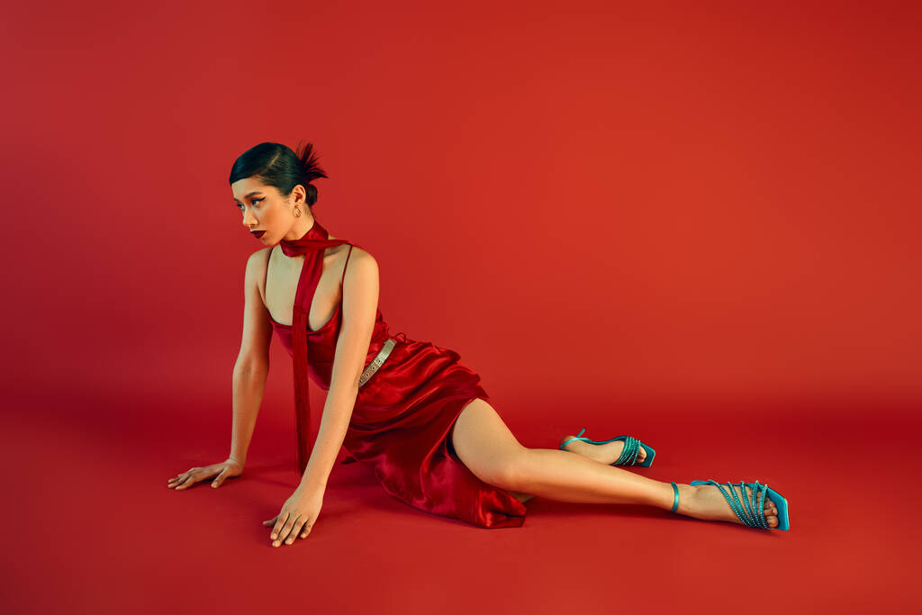 spring fashion, full length of fashionable asian woman with brunette hair and bold makeup looking away while sitting on red background in elegant strap dress, turquoise sandals and neckerchief - Photo, Image