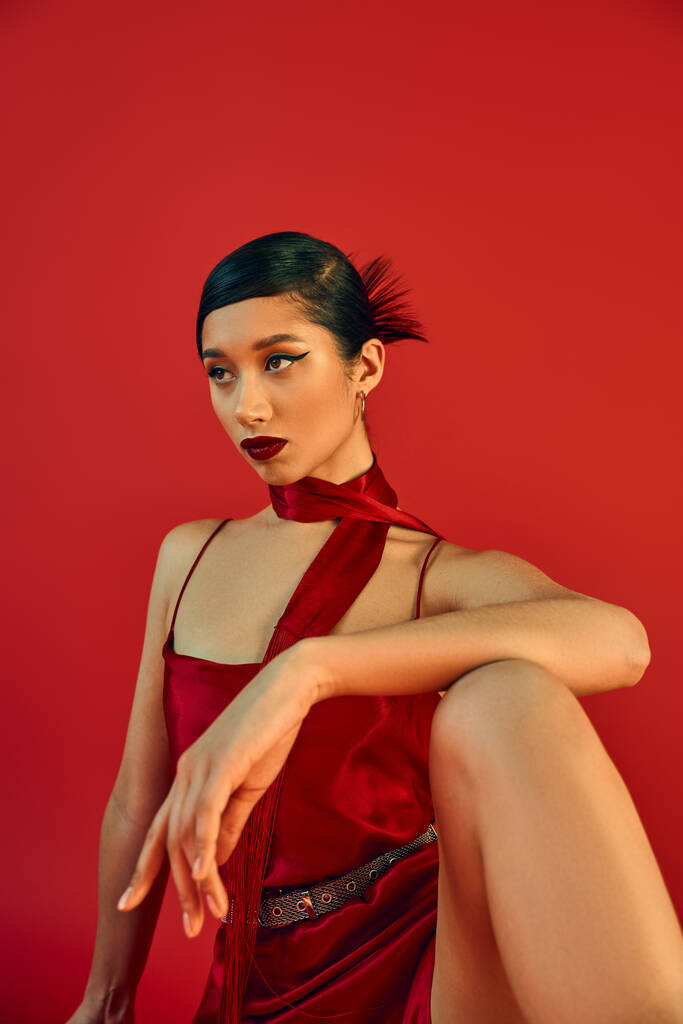 mujer asiática joven y confiada con maquillaje audaz, cabello moreno y mirada expresiva mirando hacia otro lado y posando en pañuelo y elegante vestido de correa sobre fondo rojo, fotografía de moda de primavera - Foto, imagen