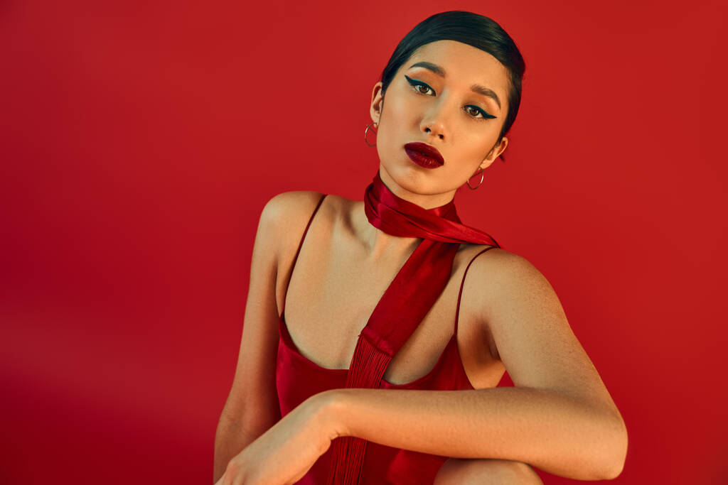 gen z mody, wiosenny styl, portret młodej i pięknej azjatyckiej kobiety z odważnym makijażem i ekspresyjnym spojrzeniem patrząc w aparat podczas pozowania w sukience pasek i chusteczkę na czerwonym tle - Zdjęcie, obraz