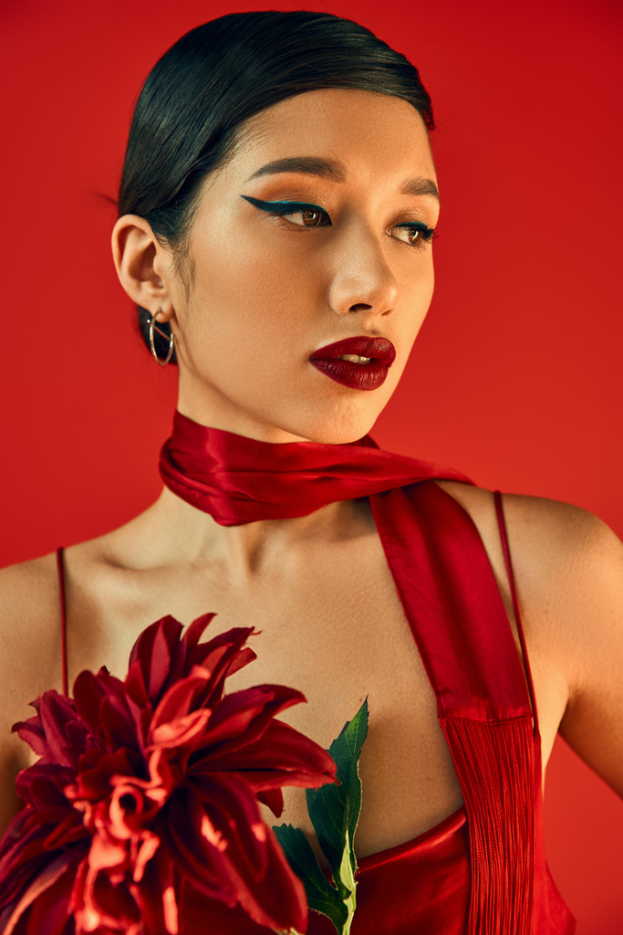 portret młodej i hipnotyzującej azjatyckiej modelki z brunetką i odważnym makijażem, pozującej w modnej chusteczce i trzymającej bordowe piwonie na czerwonym tle, koncepcja wiosennego stylu - Zdjęcie, obraz
