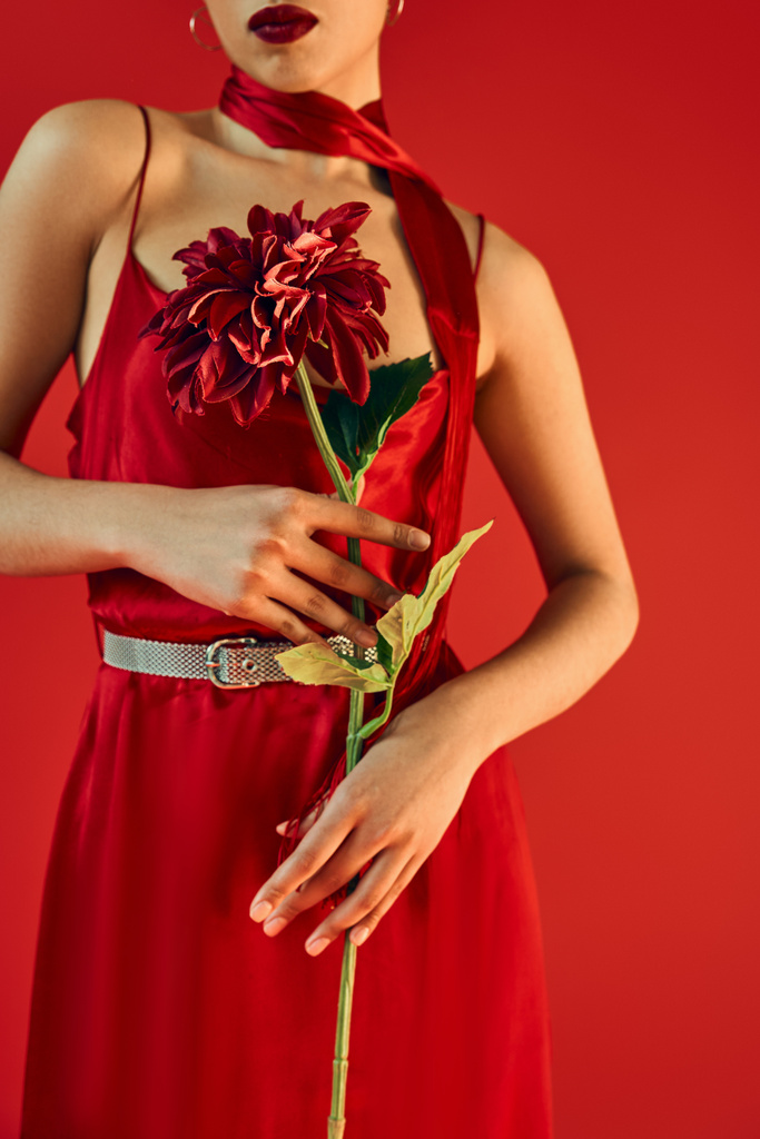 Ausgeschnittene Ansicht einer jungen Frau mit leuchtenden Lippen, in Halstuch und stilvollem Kleid mit weinroter Pfingstrose auf rotem Hintergrund, Frühlingsmodefotografie - Foto, Bild