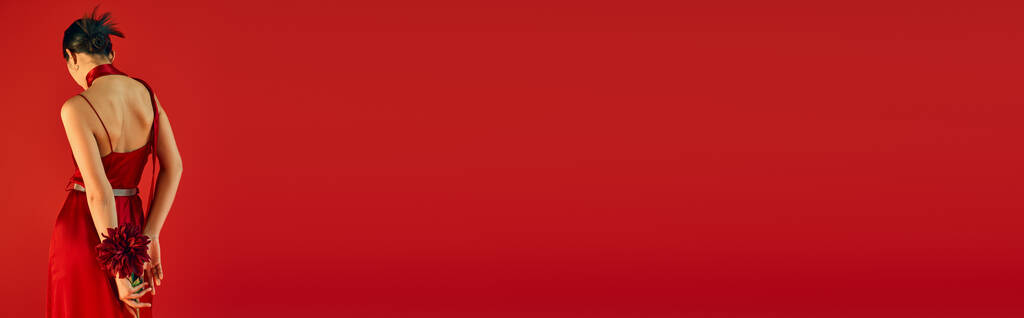 achteraanzicht van elegante en jeugdige vrouw met brunette haar houden bordeaux pioen terwijl staan in modieuze jurk op rode achtergrond met kopieerruimte, trendy lente concept, banner - Foto, afbeelding