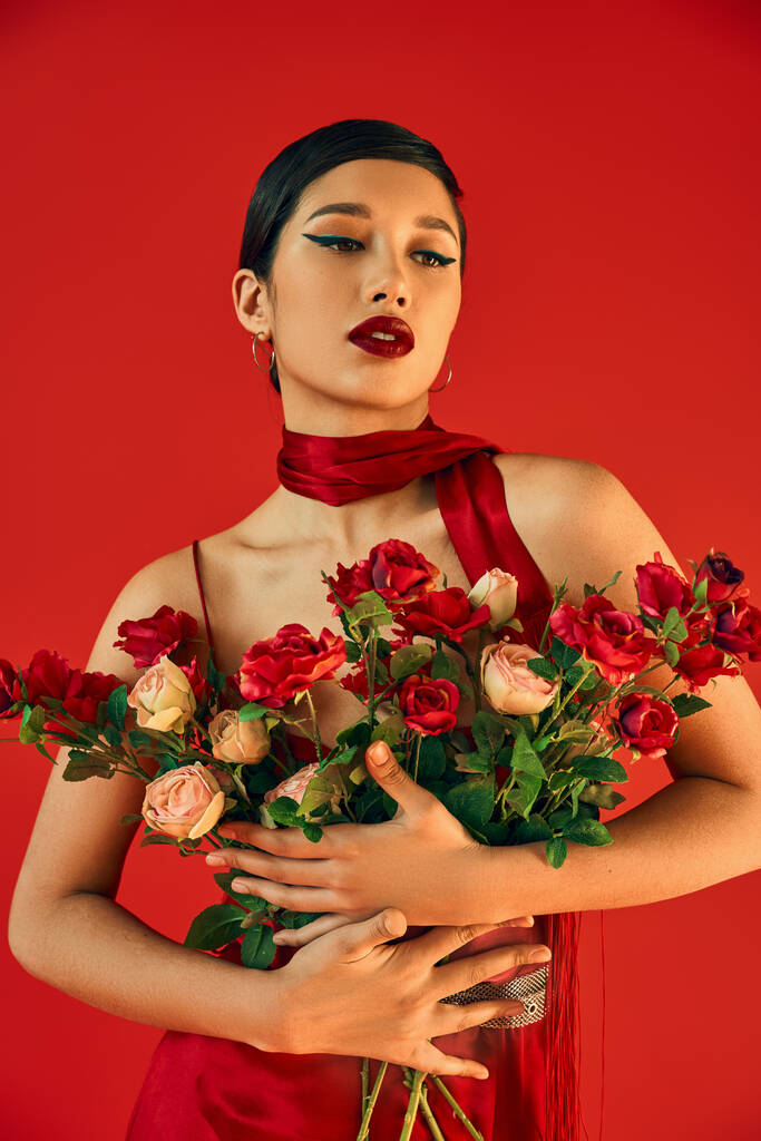 mujer asiática joven y sensual con maquillaje audaz y cabello moreno abrazando ramo de rosas mientras posando en el pañuelo sobre fondo rojo, primavera de moda, generación z - Foto, Imagen