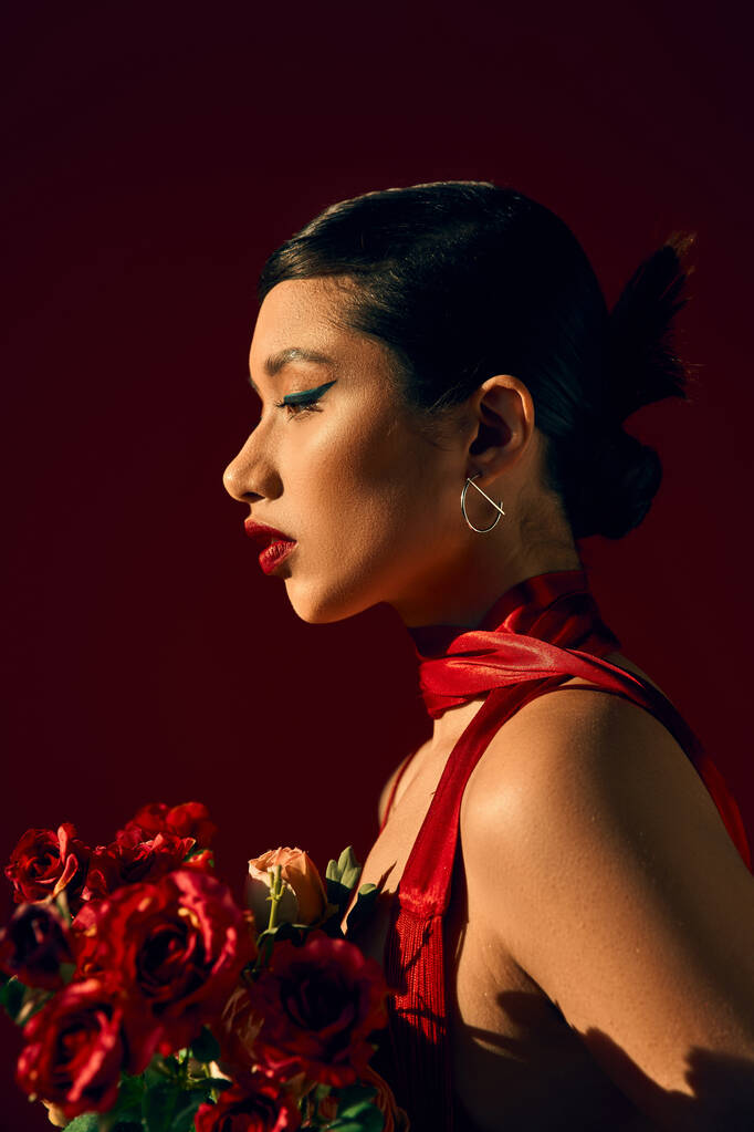 profil de charmante femme asiatique aux cheveux bruns et au maquillage audacieux, en boucle d'oreille argentée et foulard rouge tenant bouquet de roses sur fond sombre, photographie de mode de printemps - Photo, image